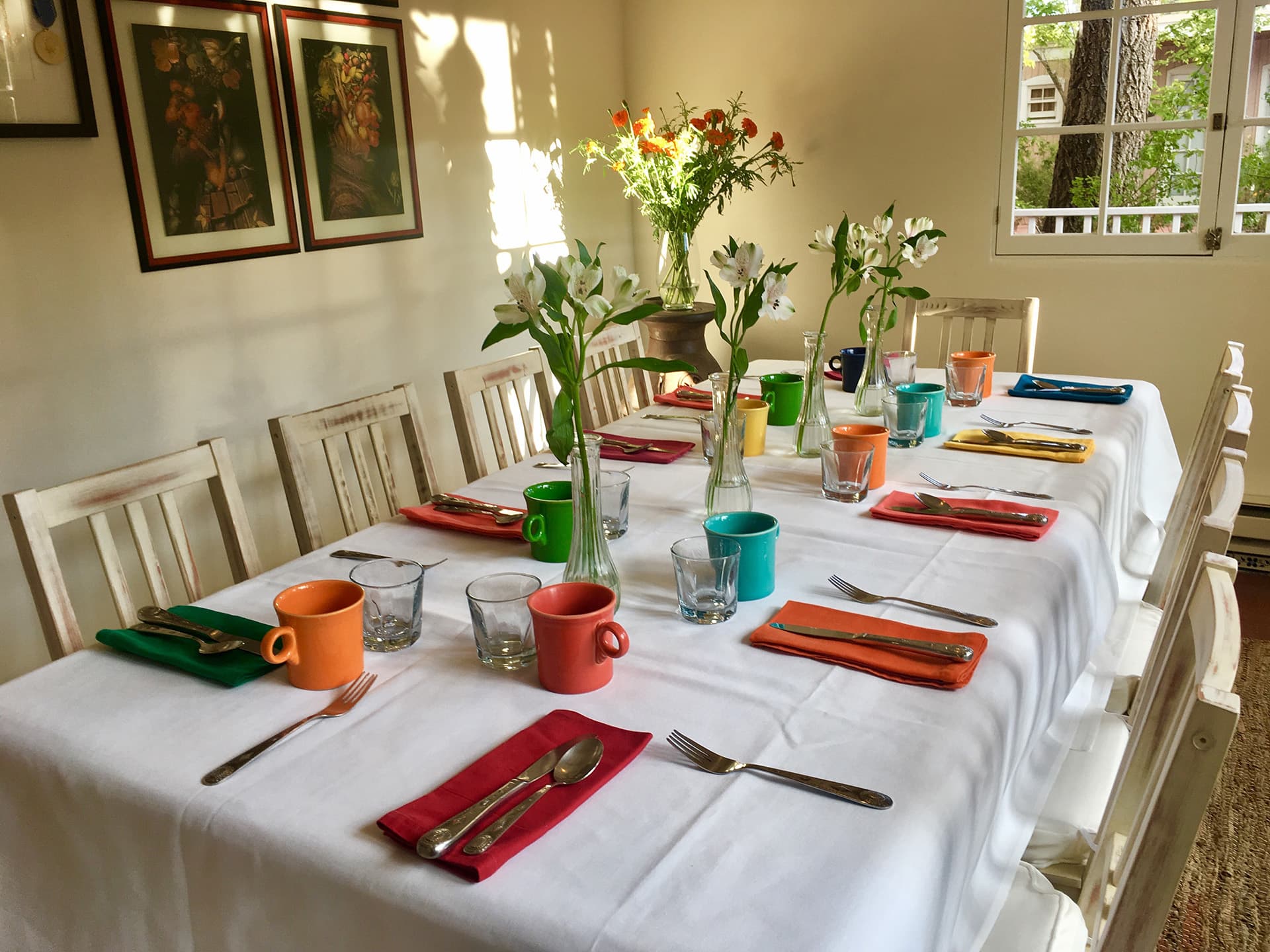 A neatly set dining table with colorful cups, vases of flowers, and silverware, bathed in warm sunlight.