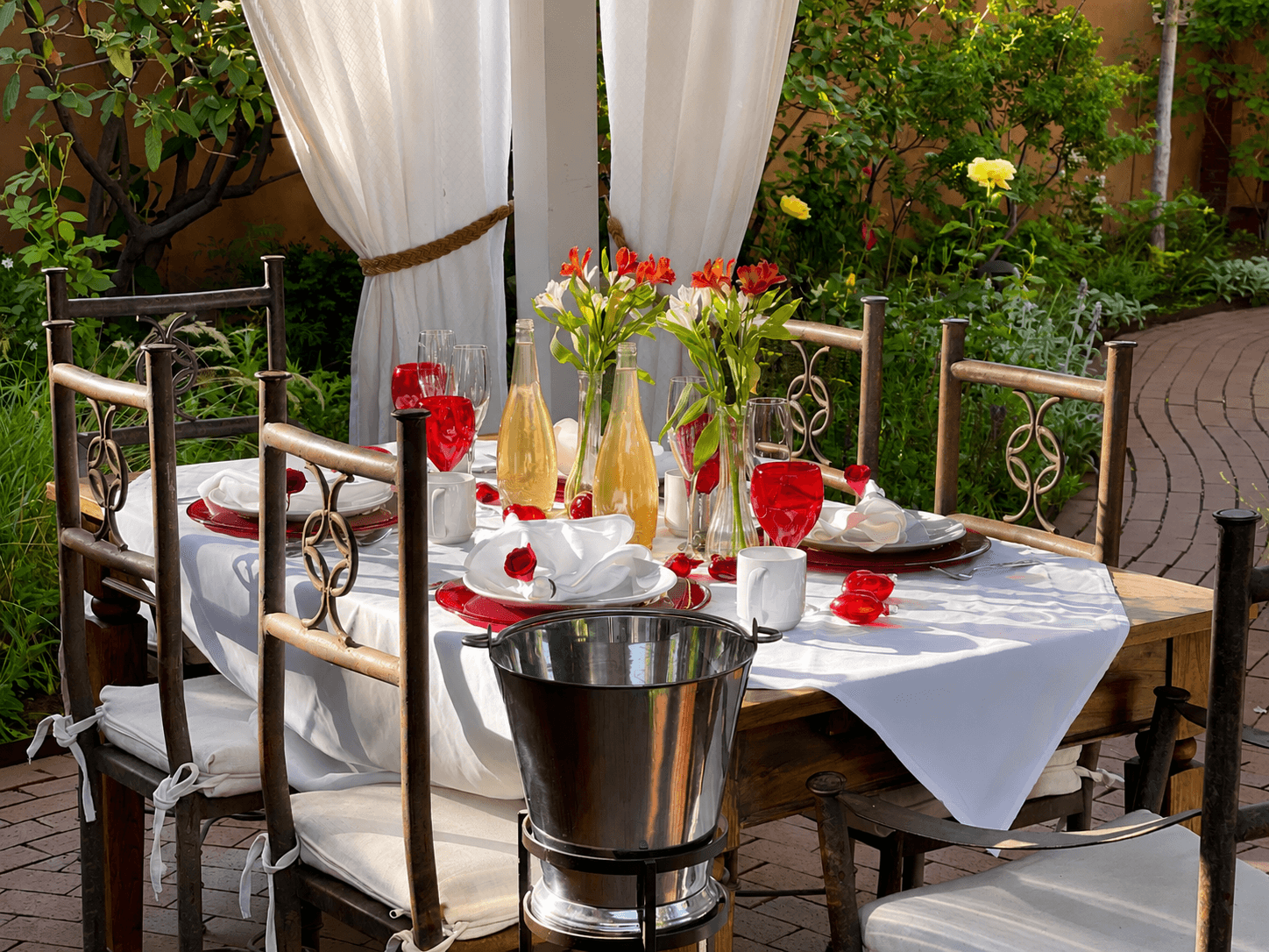 A beautifully set outdoor dining table with red drinks, flowers, and elegant tableware under a white canopy.