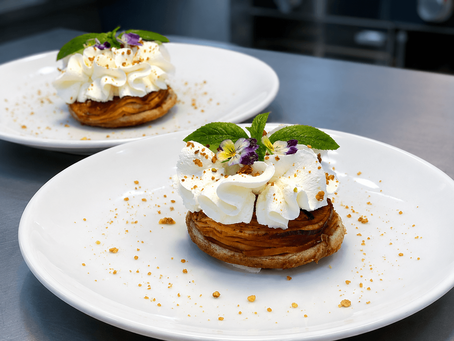 Two elegantly plated desserts topped with whipped cream, mint leaves, and edible flowers on white plates.